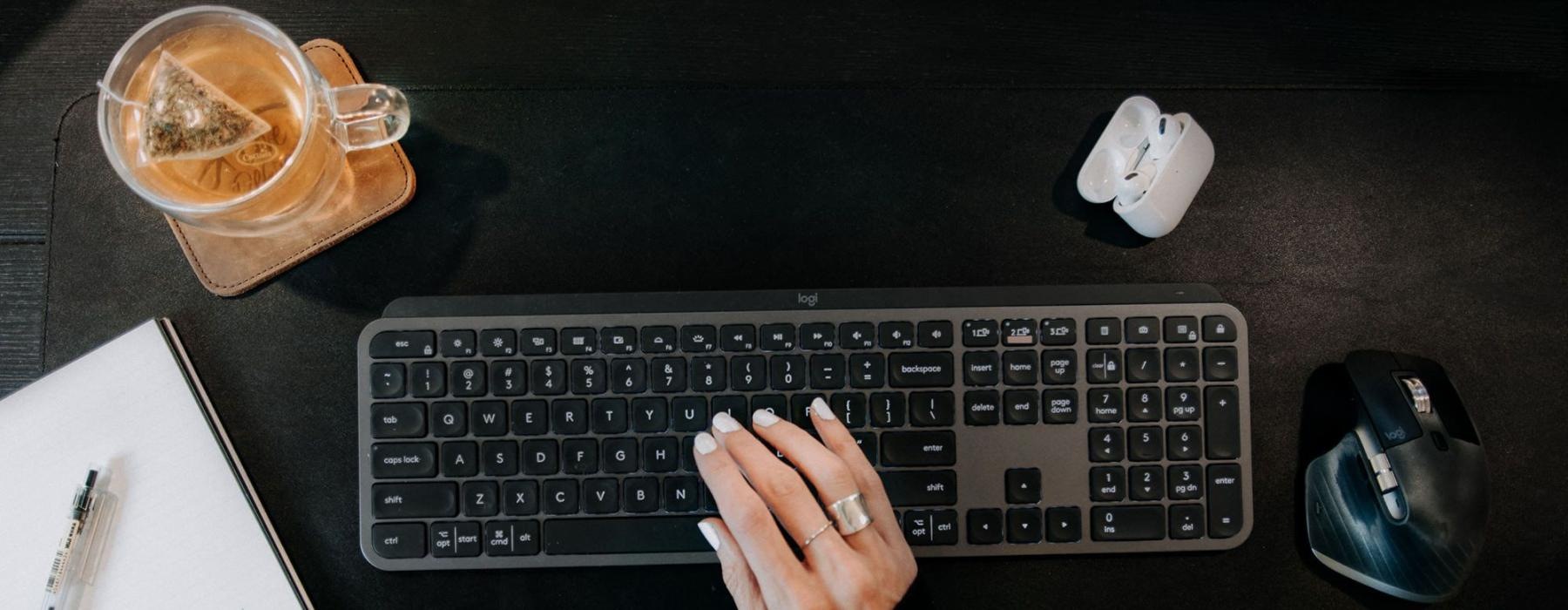 woman's hand on a keyboard surrounded by office items and a cup of tea on a coaster